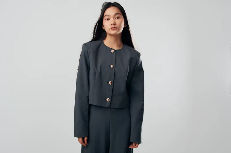 Young woman wearing dark gray suit standing against plain light background looking at camera with neutral expression. Fashion, portrait and elegance in studio for modern apparel concept.