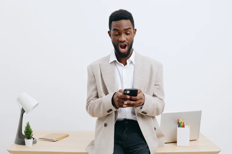 Professional young African American man in business casual attire reacting surprised while looking at his smartphone in a modern office space with minimal decor, white background, studio shot. Busines