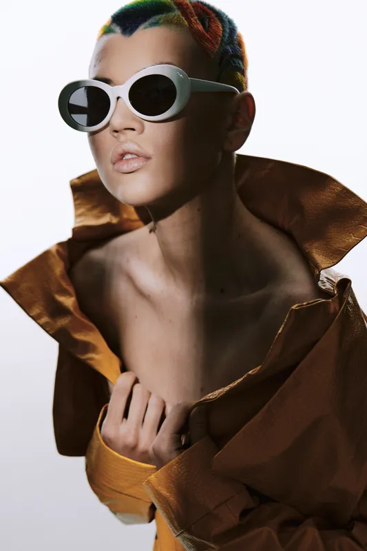 young woman with colorful hair and white sunglasses wearing a shiny brown jacket posing with confident expression in studio lighting