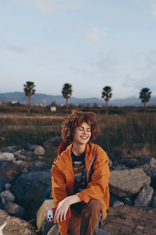 Smiling woman with curly hair in an orange jacket, sitting on rocks in an outdoor landscape, sunny daylight, palm trees and distant mountains, lifestyle travel mood, relaxed wellbeing