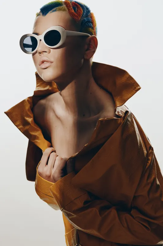 Fashion woman with colorful short hair and white sunglasses wearing a brown textured jacket posing with a confident and artistic expression on a plain background.