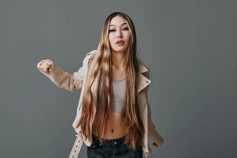 Young woman with long hair posing in casual outfit with coat and crop top against gray background. Attractive model standing with arms open and confident expression.