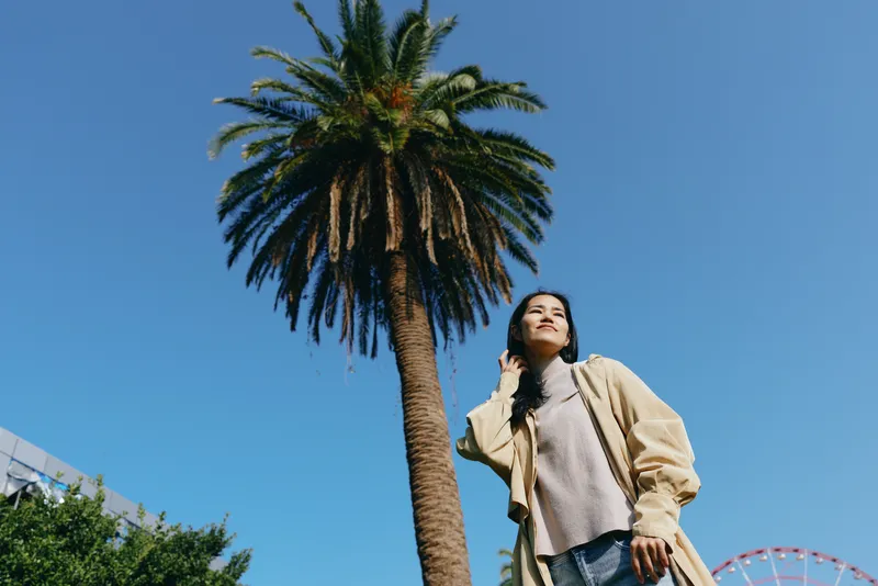 Woman under a tall palm tree in a bright outdoor setting, casual fashion, sunlight and blue sky create a relaxed lifestyle travel moment with warm breeze and vibrant greenery