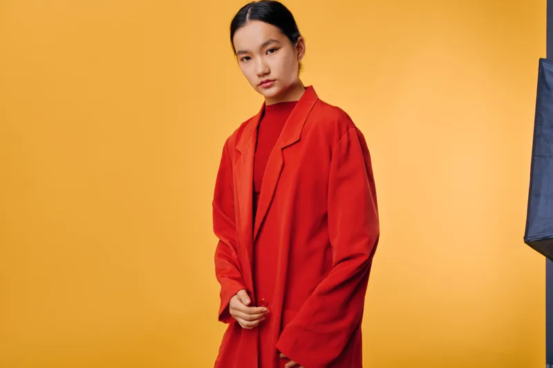 Young woman with dark hair wearing red coat and turtleneck standing against yellow background with serious expression, fashion portrait with minimal studio lighting and modern style.