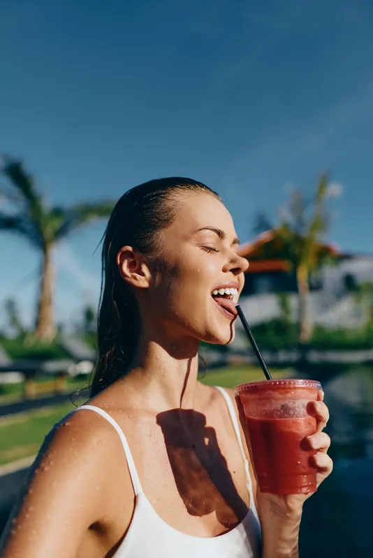Young woman in white swimsuit drinking red smoothie by the pool with palm trees in the background. Sunny day captures healthy lifestyle and summer relaxation mood.