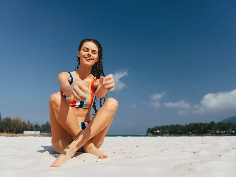 A carefree young woman sits on bright white sand at the beach, wearing a colorful striped bikini and a playful smile as the sun shines above.