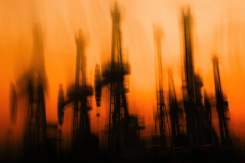 Размытое изображение нефтяных насосов на закате, демонстрирующее поразительный контраст между промышленными сооружениями и яркими оранжевыми оттенками, вызывающее ощущение энергии и индустрии