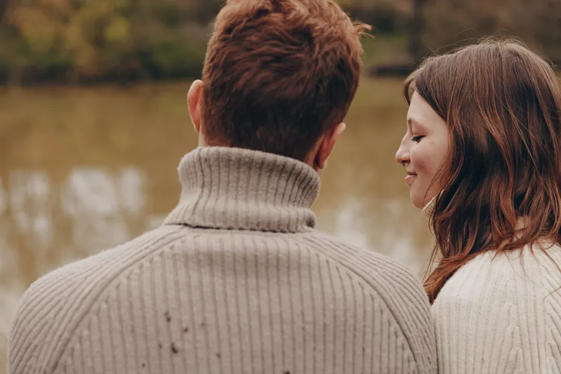 A couple stands by a calm river, seen from behind, sharing a quiet moment in cozy sweaters. Bright natural light and autumn tones create a warm, intimate mood, ideal for lifestyle and relationship