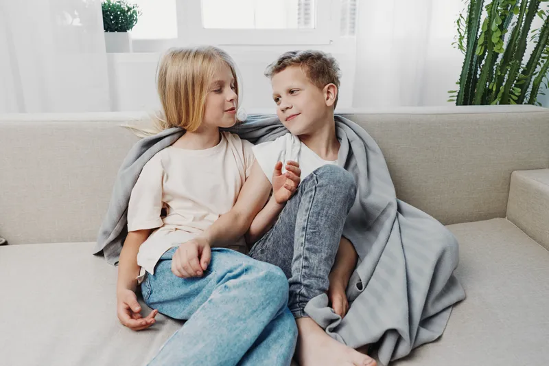 Two children sitting close on a sofa wrapped in a blanket, sharing a moment of warmth and conversation in a bright living room with natural light.
