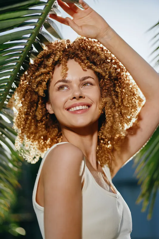 Smiling woman with curly hair stands under sunlit palm leaves, wearing white tank top and enjoying warm tropical weather with bright cheerful expression.