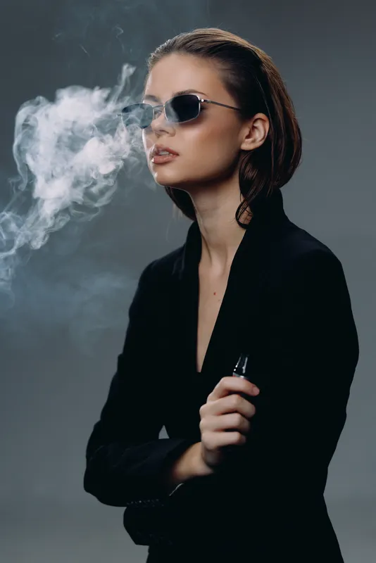Stylish woman vaping with sunglasses in dark outfit exhaling smoke with cool attitude