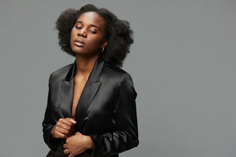 Elegant young woman in a black satin blazer with natural hair, exuding confidence and sophistication against a plain gray background