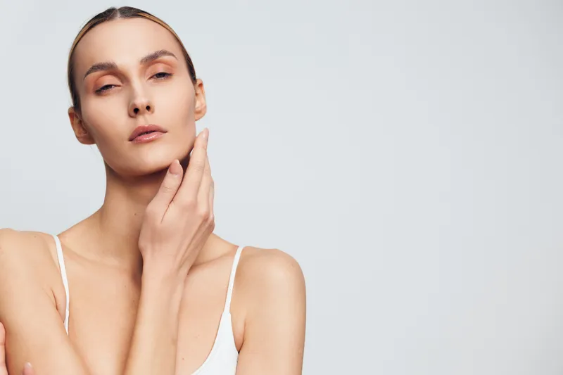 A young woman with clear skin touches her face gently while looking at the camera. The minimalistic background emphasizes natural beauty and skincare concept.