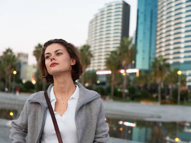 A confident professional woman stands outdoors in a modern cityscape, looking upward with a thoughtful expression. The soft daylight highlights her poised demeanor and ambition.