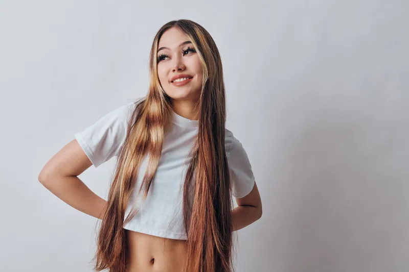 Young woman with long braided hair standing against white wall with hands behind back smiling and looking sideways in casual white crop top and relaxed pose.