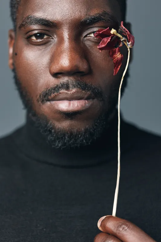 African man with dark skin holding dried red flower near eye in creative conceptual visual style. Portrait closeup with black turtleneck and serious expression on gray background.