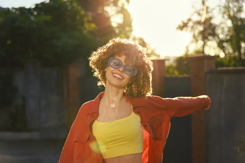 Young woman with curly hair dances in a sunlit outdoor space, wearing a yellow top and orange jacket, radiating carefree confidence and bright energy