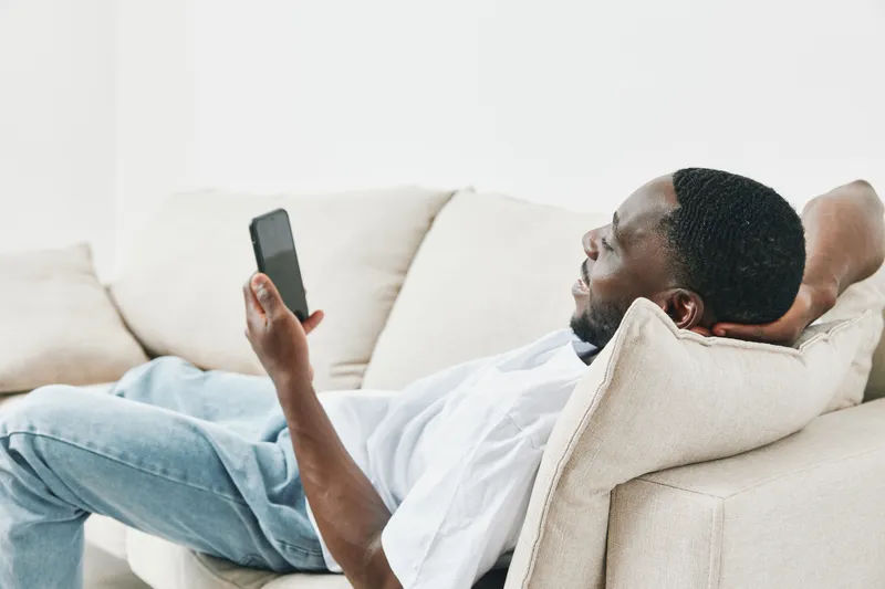 Relaxed young African man lounging on a beige couch while browsing on a smartphone, capturing a moment of leisure and modern life in a bright, minimalistic setting