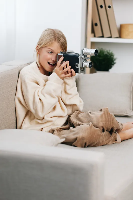 Authentic lifestyle scene featuring a playful student girl with a vintage camera, capturing candid moments on a cozy sofa in a sunlit living room, inviting warmth.