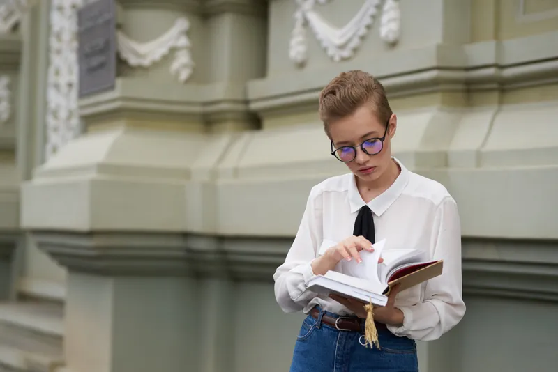Счастливая студентка стоит возле здания с книгой в руке и в очках на лице