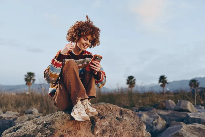 Woman lifestyle outdoors, smiling relaxed with curly hair, holding a phone on a rock, striped sweater, natural light, open air, soft breeze, palm trees in the distance, cloudy sky