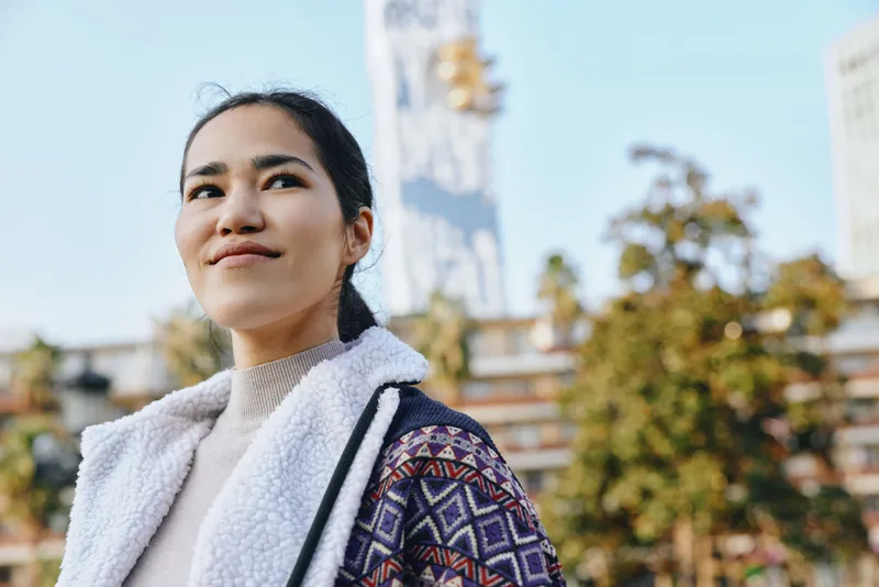 Outdoor image of a smiling woman wearing a cozy patterned jacket and fleece, standing in a sunny urban park with trees and buildings behind, conveying confidence, style, and modern lifestyle