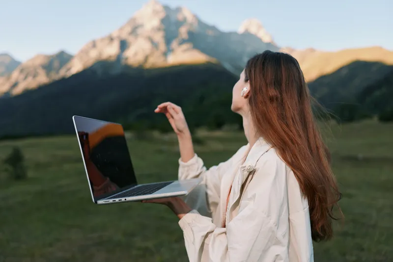 A cheerful woman uses a laptop outdoors in a scenic landscape. She smiles and types with focus, creating a positive and productive mood in a fresh, natural setting.
