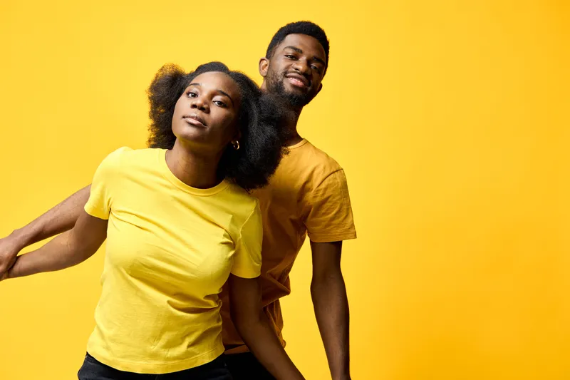 Young African American couple joyfully posing together on a yellow background, showcasing vibrant clothing and playful emotions