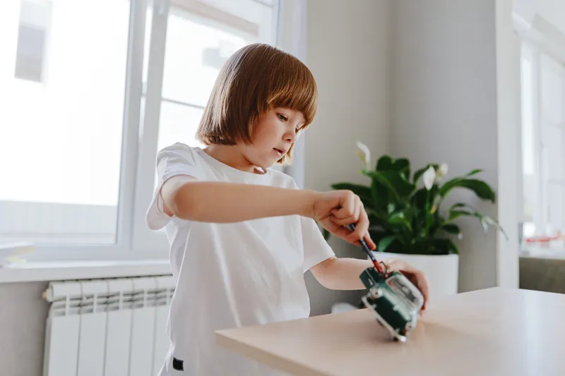 Curious young boy engaging with a toy robot, using a screwdriver in a bright and cheerful indoor environment, showcasing creativity and exploration.