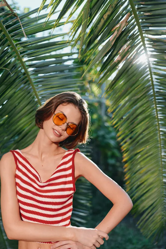 A cheerful woman enjoys a sunny outdoor setting beneath palm leaves. She wears a red striped top and round sunglasses, smiling warmly and radiating a relaxed vacation vibe.
