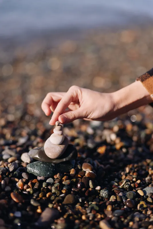 Stone balancing on a beach with a hand placing smooth rocks into a small stack among pebbles and sand, calm focus and meditation in a natural outdoor setting