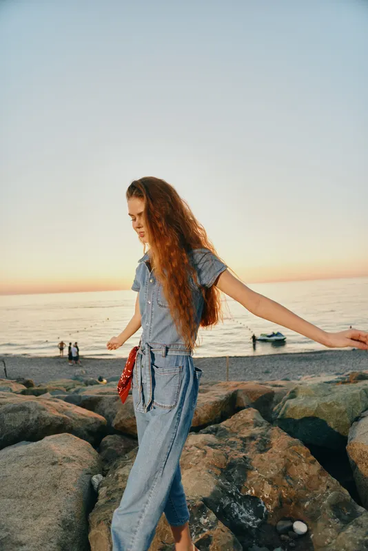lifestyle woman in modern boho-western denim street style walking on rocks by the sea at sunset with film color warmth and natural light