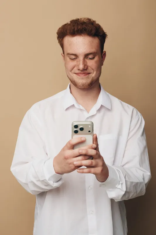 Smiling young man with curly hair wearing white shirt looks at smartphone with closed eyes. Neutral beige background. Concept of technology, communication, happiness, and casual lifestyle.
