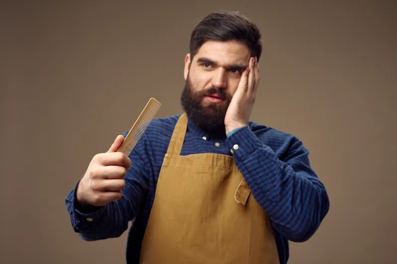 Мужчина с бородой касается рукой своего лица и расчёски на бежевом фоне парикмахерской, одетый в фартук