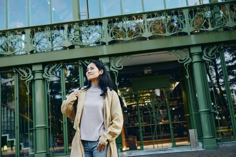 Woman wearing a beige coat stands outdoors in front of a glass storefront, showcasing urban style and relaxed confidence in cool weather and sunlight