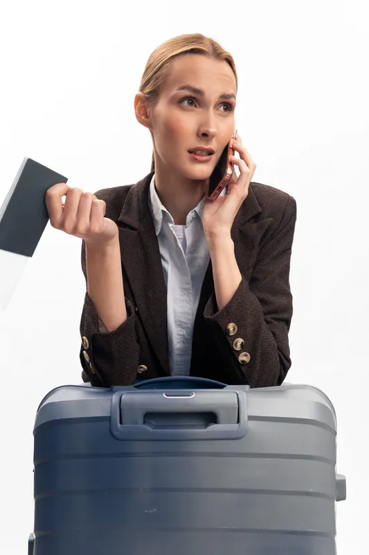 Businesswoman with suitcase holds passport talking on phone, looking worried or concerned. Travel concept with stress, urgency, and last minute issues in white studio background.