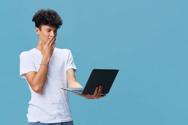 Young man with curly hair holding laptop, wearing casual white t-shirt, thoughtful expression, standing against solid blue background, technology and concentration concept.