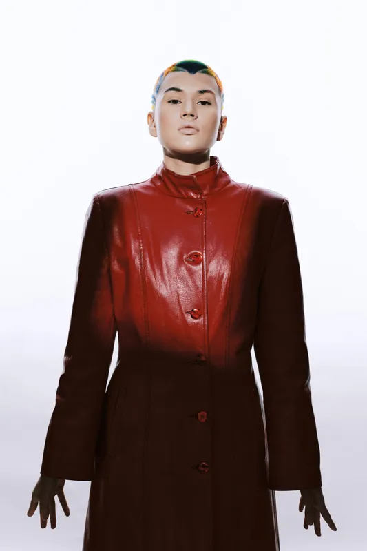 Young woman with short hair wearing a red leather coat, standing against a bright white background with a serious expression and modern fashion style.