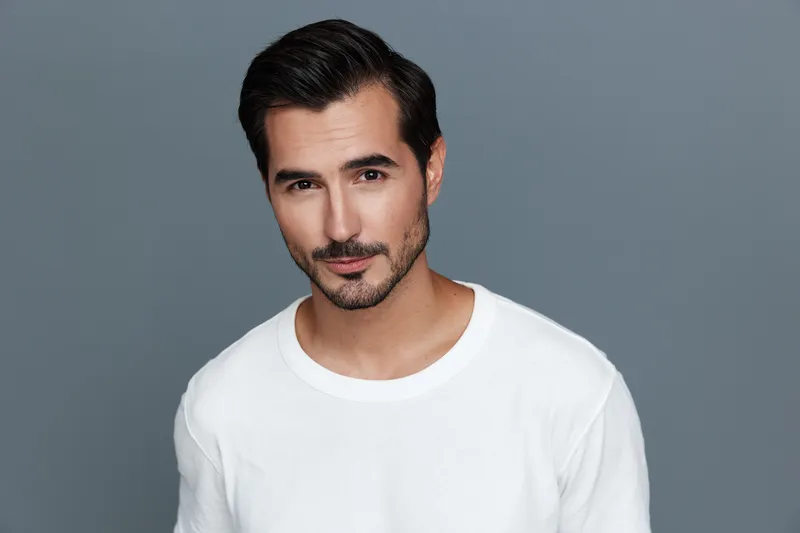 Confident handsome man in white shirt, smiling at camera, posing against solid grey background. Stylish portrait reflects modern lifestyle and self-assurance.