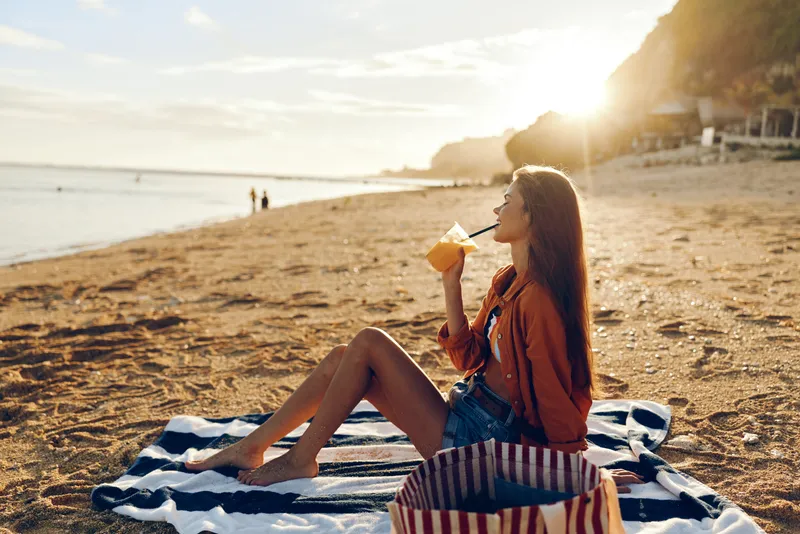 Woman beach sunset relaxation, sitting on blanket, young girl reads a book, calm ocean shoreline behind, golden sunlight on sand, summer vacation getaway, peaceful evening ambience