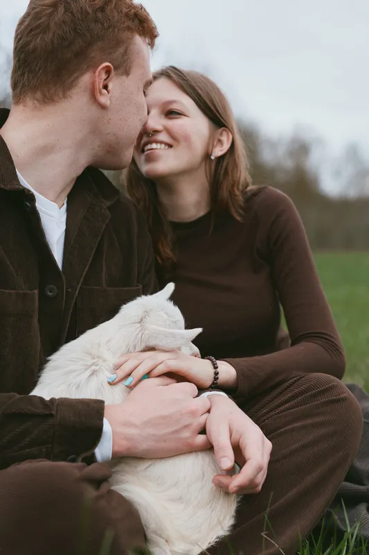 Smiling couple sharing a kiss while holding a goat kid in a grassy field, capturing affection, outdoors lifestyle, romance and nature in a candid moment with animals.