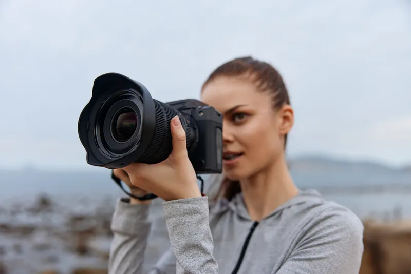 A focused young woman photographing a coastal landscape, dressed in a casual grey hoodie, showcasing determination and creativity