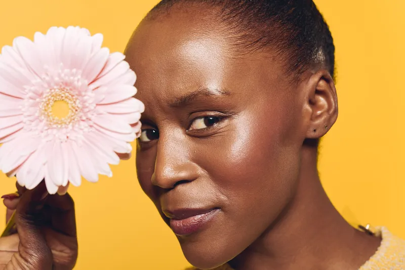 Close-up portrait of a smiling African woman with dark skin holding a pink flower near her face. Bright yellow background, natural beauty, skincare, wellness, and floral concept.