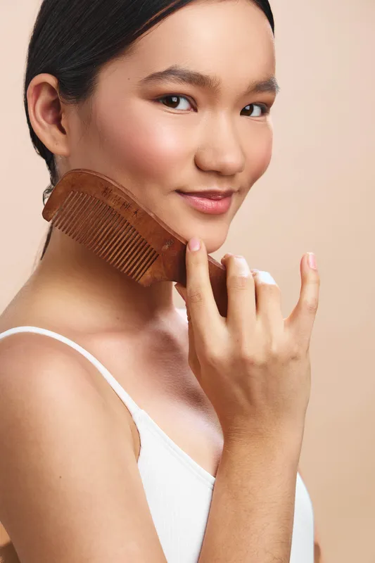 asian woman in close up with a wooden comb near her face, warm studio light, soft smile, presenting a calm selfcare moment and contemporary grooming scene for lifestyle and beauty projects.