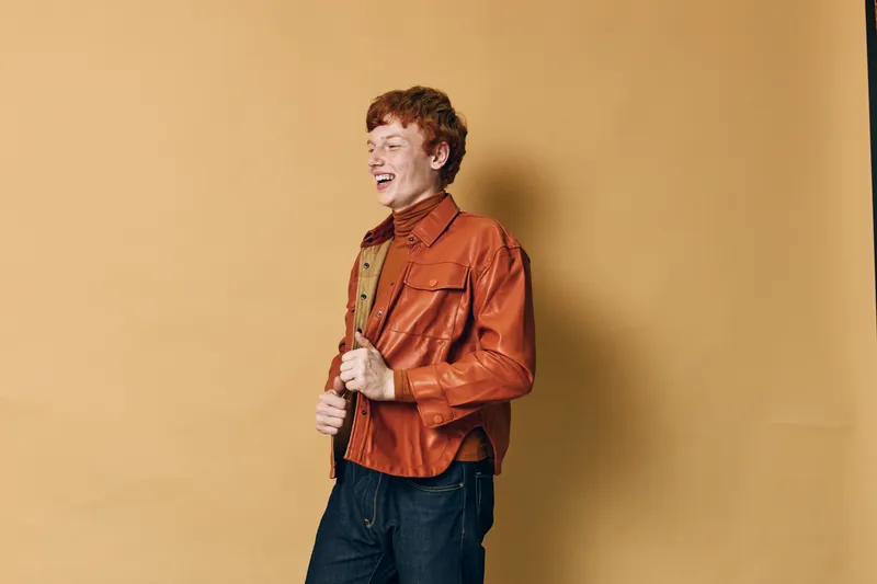 Young man with red hair wearing brown leather jacket and dark jeans, smiling and standing against beige background in casual outfit for fashion style.