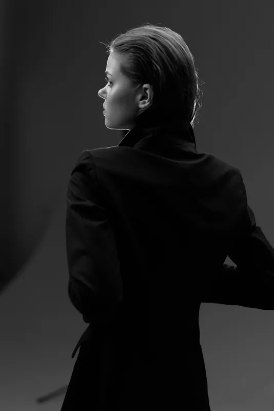 Black and white portrait of a stylish woman in a dark coat looking away with elegant posture and moody lighting