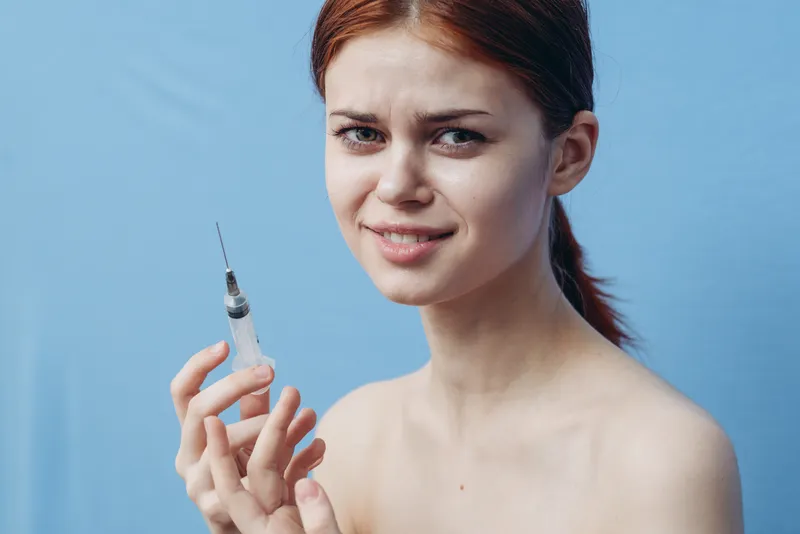 Женщина держит шприц возле лица, делая инъекцию для омоложения ботоксом, на синем фоне