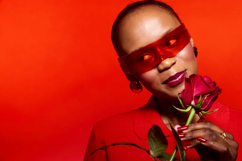 Elegant black woman with dark skin wearing red glasses and red blazer holds a red rose close to her face. Studio portrait on red background. Concept of fashion, beauty, and style.
