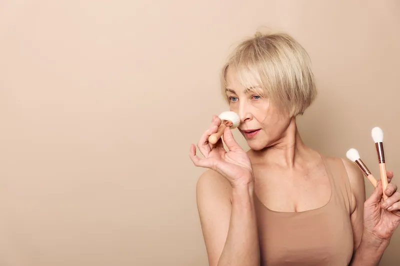 Mature woman with short blonde hair applies makeup using soft brushes on neutral background. Concept of beauty routine, natural skincare, and self-care for older women in minimalist style.