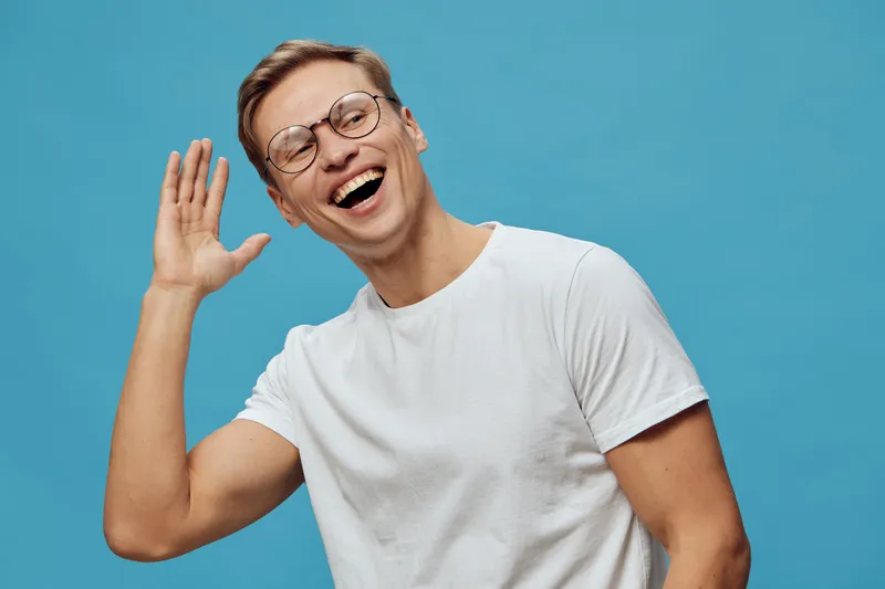 Smiling young man with short hair and glasses wearing casual white t-shirt, happy cheerful expression, playful pose on plain blue studio background, people lifestyle concept